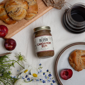 A jar of Cardamom Bun Almond Butter sits on a white floral tablecloth surrounded by plums, flowers, a carafe of coffee, cardamom buns, and swedish flatbreads.