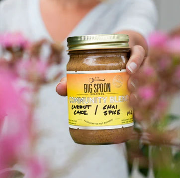 A person holds a jar of Big Spoon Roasters Community Blend nut butter. Pink flowers are visible in the background.