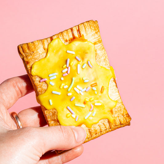 A hand holds a pastry tart with yellow frosting and rainbow sprinkles in front of a pink background