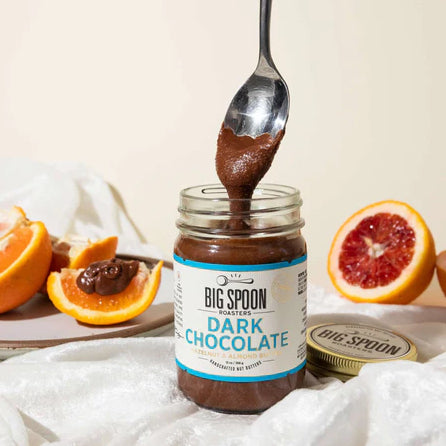 A spoon pulls Dark Chocolate Hazelnut & Almond Butter from an open jar on a cream-colored background. Orange and Blood Orange halves are visible in the background.