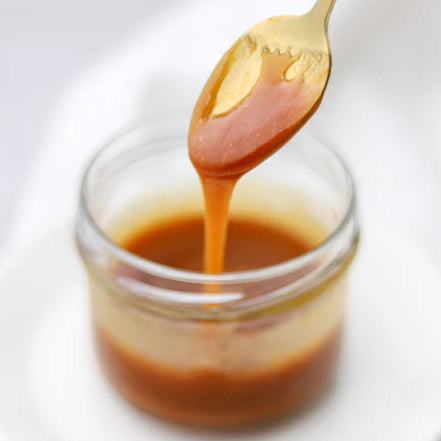 Caramel Sauce in a small glass jar with a gold spoon pulling some caramel out on top of a white saucer and white background.