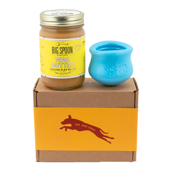 Jar of Banana Flax Wag Butter and a blue West Paw Toppl toy sit on top of a gift box with a gold and maroon decorative dog label.