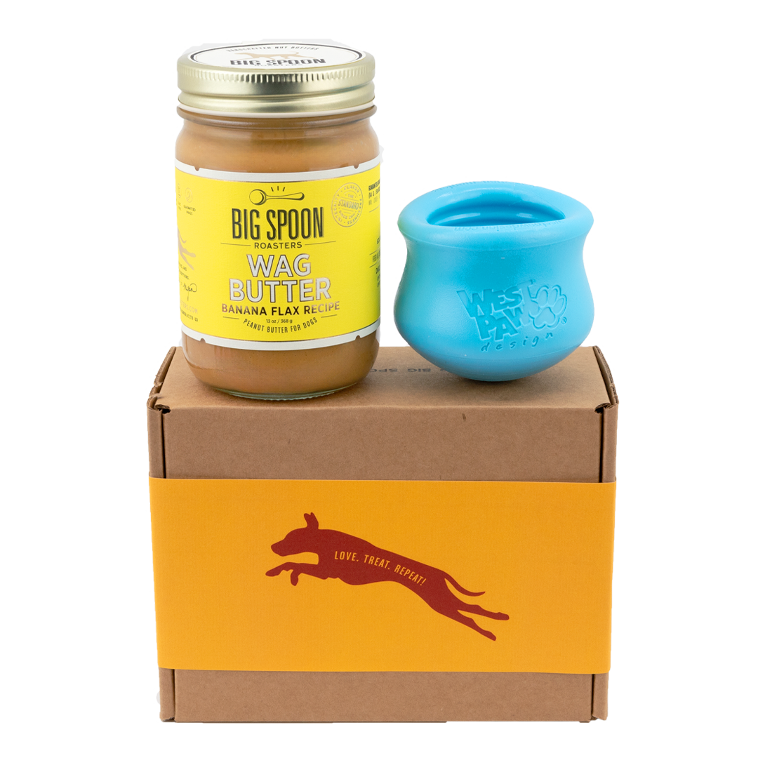Jar of Banana Flax Wag Butter and a blue West Paw Toppl toy sit on top of a gift box with a gold and maroon decorative dog label.