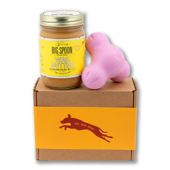 Jar of Banana Flax Wag Butter and a pink West Paw Tux toy sit on top of a gift box with a gold and maroon decorative dog label.
