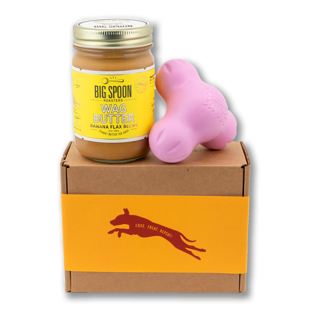 Jar of Banana Flax Wag Butter and a pink West Paw Tux toy sit on top of a gift box with a gold and maroon decorative dog label.