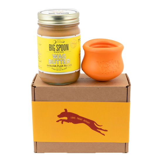 Jar of Banana Flax Wag Butter and a orange West Paw Toppl toy sit on top of a gift box with a gold and maroon decorative dog label.