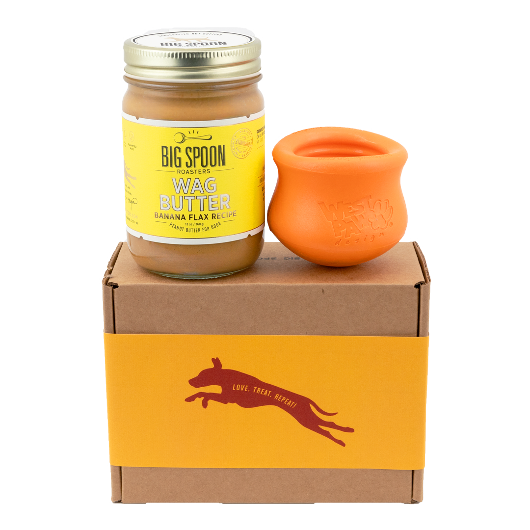 Jar of Banana Flax Wag Butter and a orange West Paw Toppl toy sit on top of a gift box with a gold and maroon decorative dog label.