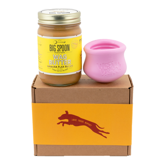 Jar of Banana Flax Wag Butter and a pink West Paw Toppl toy sit on top of a gift box with a gold and maroon decorative dog label.