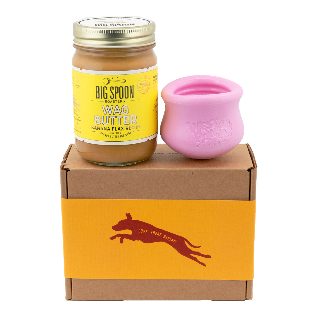 Jar of Banana Flax Wag Butter and a pink West Paw Toppl toy sit on top of a gift box with a gold and maroon decorative dog label.