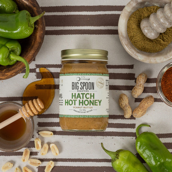 Jar of Hatch Hot Honey Peanut Butter surrounded by peanuts, hatch chiles, and honey