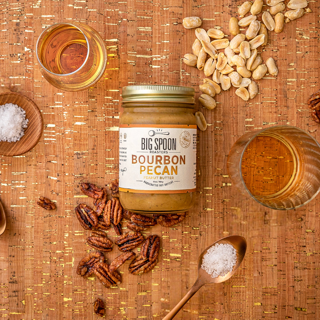 A 13oz jar of Bourbon Pecan surrounded by pecans, peanuts, sea salt, maple syrup, and bourbon with a cork and golden background