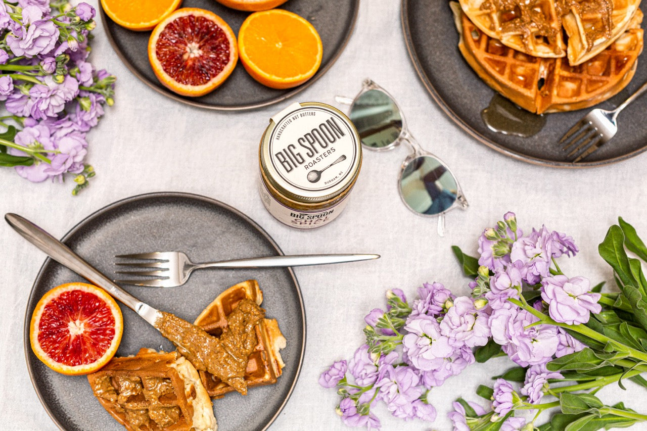 A jar of big spoon roasters nut butter surrounded by plates of waffles spread with nut butter, fresh fruit, and flowers