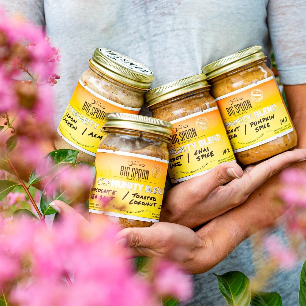 Hands holding four jars of Community Blend nut butter with pink flowers around it