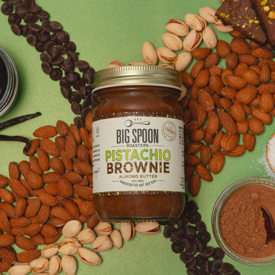 Jar of Big Spoon Pistachio Brownie Nut Butter surrounded by almonds, pistachios, chocolate chips, vanilla, coconut nectar, cocoa powder, a spoonful of salt, and pistachio-topped brownies on a green background.