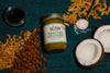 A jar of Toasted Coconut Almond Butter sits alongside almonds, toasted coconut chips, a jar of coconut nectar, a spoonful of sea salt, and a whole opened coconut