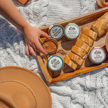 A hand reaches for a 3oz jar of nut butter surrounded by other 3oz nut butter jars, a sliced baguette, and a caramel hat all on top of a cable knit white picnic blanket