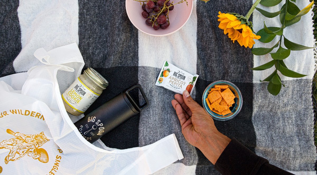 A shopable picnic spread with a hand reaching for an Apricot Pepita bar surrounded by a Big Spoon Roasters tote bag, water bottle, Lemon Cookie nut butter along with fresh fruit and flowers on a black and white checkered blanket