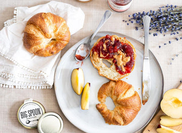 A halved croissant is spread with peanut butter and jam surrounded by fresh fruit, croissants, and lavender sprigs on a linen table cloth