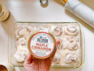 A hand holds a 3oz jar of Maple Cinnamon nut butter in front of a batch of cinnamon rolls