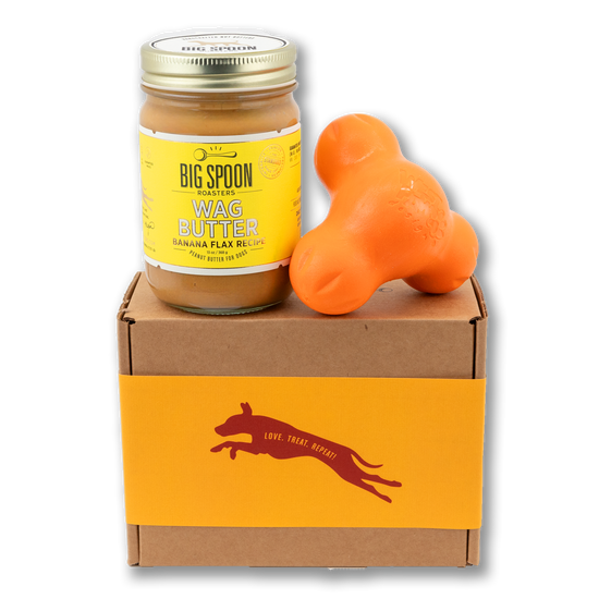 Jar of Banana Flax Wag Butter and an orange West Paw Tux toy sit on top of a gift box with a gold and maroon decorative dog label.