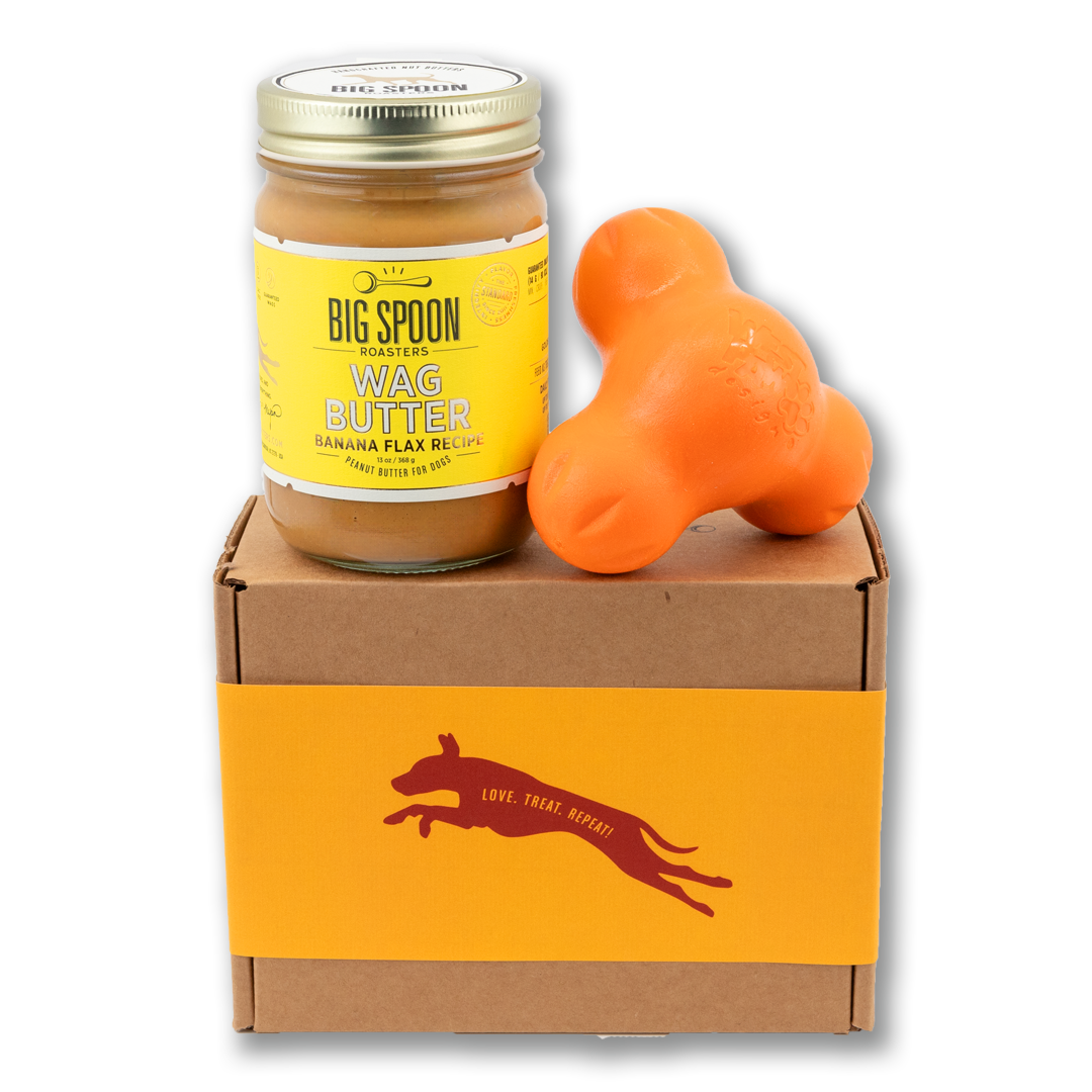 Jar of Banana Flax Wag Butter and an orange West Paw Tux toy sit on top of a gift box with a gold and maroon decorative dog label.