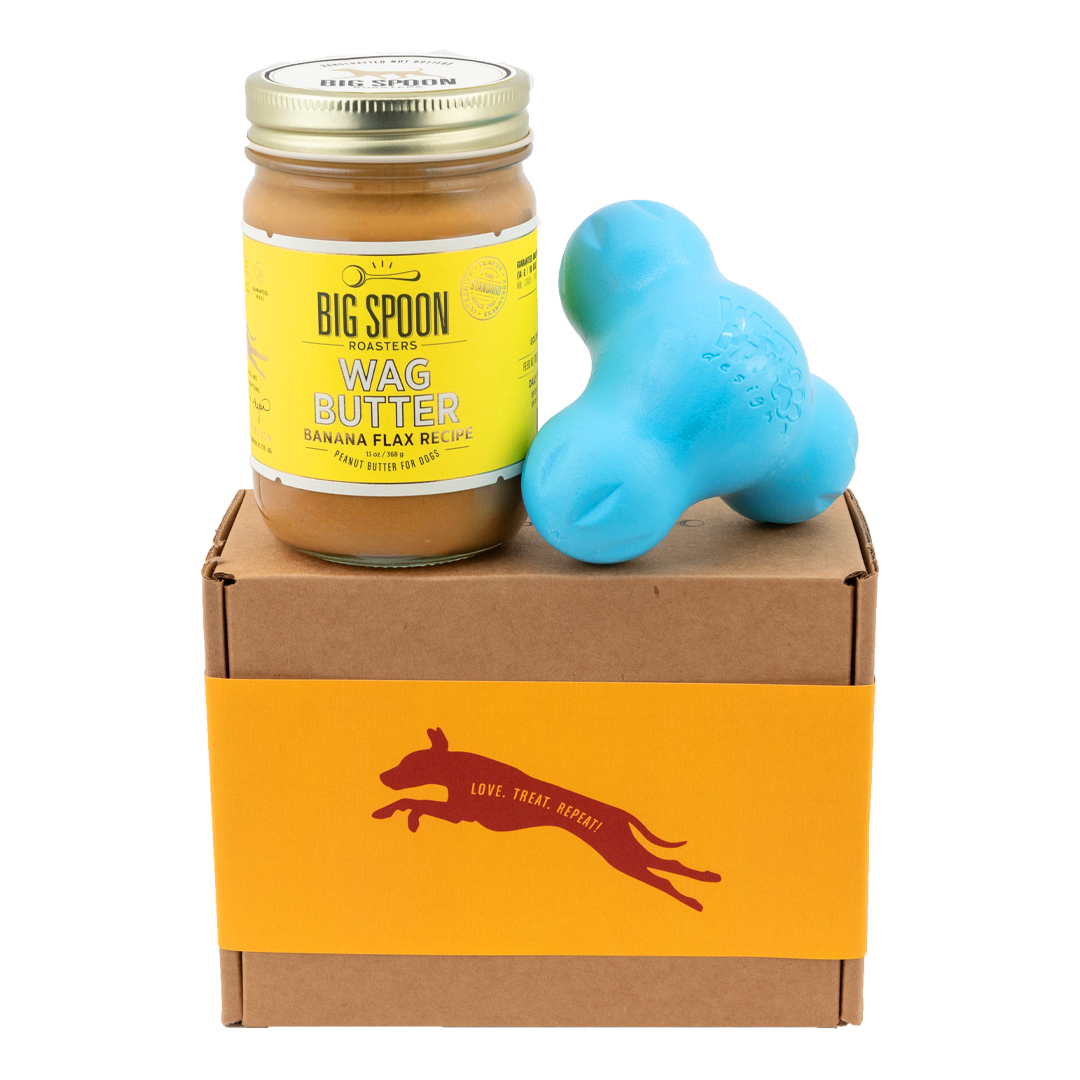 Jar of Banana Flax Wag Butter and a blue West Paw Tux toy sit on top of a gift box with a gold and maroon decorative dog label.
