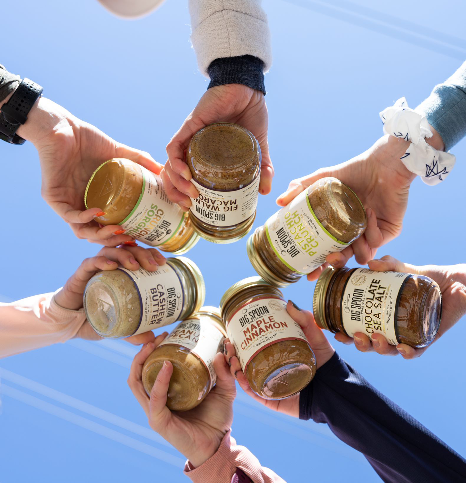 Seven hands, each holding a jar of nut butter.